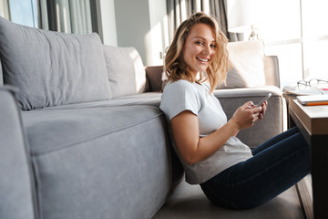 Image of happy woman smiling and using cellphone while sitting on floor
