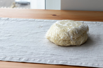Raw curd dough with bits of cottage cheese is wrapped in plastic wrap on a linen tablecloth on a wooden table. The process of making curd bagels or croissants