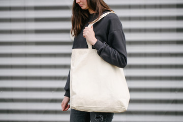 Young woman holding white textile eco bag against urban city gray background outside wearing black...