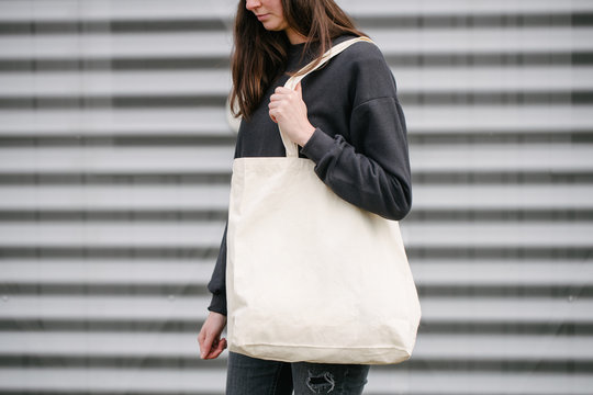 Young woman holding white textile eco bag against urban city gray background outside wearing black casual clothes. Ecology or environment protection concept. White eco bag for mock up