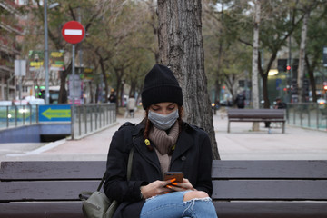 Obraz premium Young woman sitting on a street bench wearing medical mask and looking at her smartphone