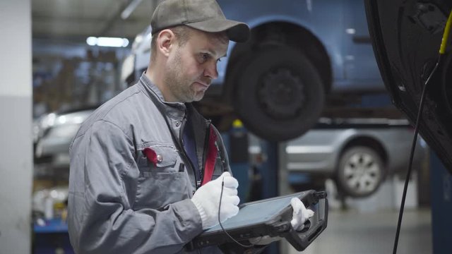 Side view of confident Caucasian man in workwear entering data into computer in repair shop. Adult professional auto mechanic fixing automobiles at car service station. Job, occupation, lifestyle.