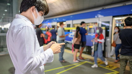 Fototapeta na wymiar Asian man wearing surgical face mask using smartphone at skytrain station platform. Wuhan coronavirus (COVID-19) outbreak prevention in public transportation. Health awareness for pandemic protection