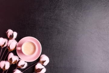 Greeting card from pink ceramic cup with fresh coffee drink and natural cotton flowers on a black eco leather background.