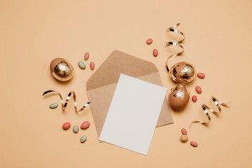 Blank craft envelope with a read sheet of paper, gold glitter tape on a beige background. Golden easter eggs with jelly beans candy, top view.