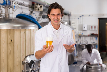 Young man brewer in uniform is standing with glass of beer