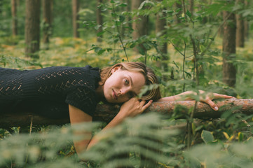 Portrait of young beautiful girl in black dress in forest that lies on the tree trunk - 327875014