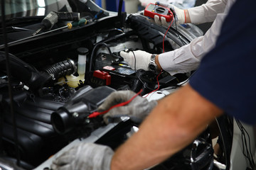 Close-up view of professional garage mechanics working with multimeter or voltmeter checking a car battery level. Open trunk with car details. Service center concept