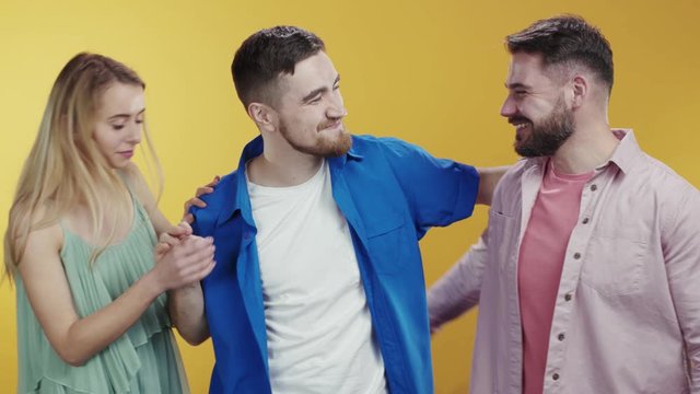 Group of caucasian young friends with girl posing on orange background. Two best friends men kick out girl embracing each other brothers smile happy at camera. Break up, Relationships.