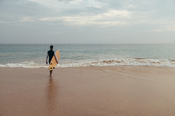 Handsome teenage boy with surfboard going into the ocean at sandy beach on surf line.
