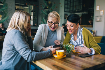 mid aged woman friends talking and drinking coffee in cafe, using mobile phone