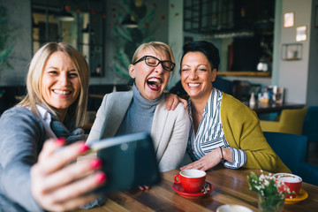 mid aged woman friends taking picture with mobile phone while drinking coffee in cafe