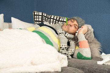 thickly dressed man with a thermometer is lying on his couch with the flu