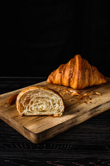 A croissant cut in half and a whole one on a wooden board, crumbs around, on black background