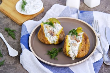 Whole baked potatoes with sour cream, herring and pickled cucumbers sauce on a brown plate on a gray concrete background. Potato recipes