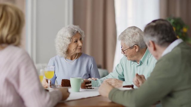 Group of four senior friends sitting at dining table at home and enjoying talk over dessert. Smiling elderly woman with curly grey hair speaking