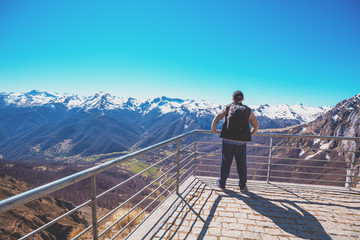 A man tourist stands on a viewpoint above valley against mountains covered with snow. A man looks...