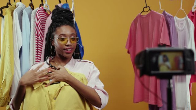 Happy african american trendy girl vlogger filming live fashion vlog streaming showing stylish clothes items isolated on background. Fashion influencer. Try-on haul.