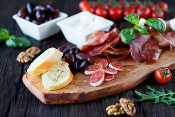 Prosciutto, bread, olives, walnut, mozzarella, salami, basil and cherry tomatoes on  brown wooden board.  Mediterranean kitchen.
