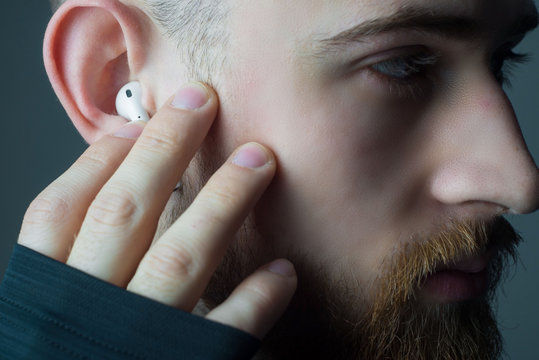 Close-up portrait of a young bearded guy of twenty-five years old, looking at the camera, in wireless white headphones in his ears. Studio advertising photo of headphones. Athlete in headphones
