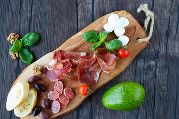Prosciutto, bread, olives, walnut, mozzarella, salami, basil, avocado and cherry tomatoes on  brown wooden board.  Mediterranean kitchen.