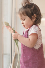 A girl in a pink sundress holds a tulip. A child is standing at the window.