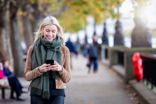 Eine attraktive, blonde Frau läuft auf der Straße und liest ihre Emails und Nachrichten auf dem Mobiltelefon