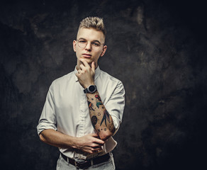 Young student guy in a white shirt, with tattoos on his hand, standing in a thinking pose, wearing glasses and watches, in a dark studio isolated on grey background, looking stylish and thoughtful