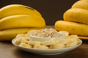 A bunch of bananas and a sliced banana on a plate with oats on a table