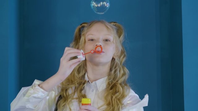 Cute girl blowing soap bubbles. Slow motion view of curly blonde girl inflating colorful soap bubbles. Leisure concept
