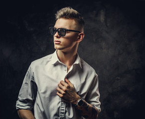 Cute and inked hipster male model posing in a studio wearing a white shirt, sunglasses, standing in front of the grey background, looking cool and interested