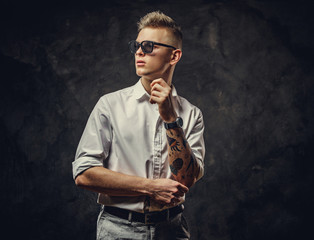 Fototapeta na wymiar Extravagant and inked hipster male model posing in a studio wearing a white shirt, sunglasses, standing in a cool pose in front of the grey background, looking interested