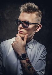 Young handsome adult student guy in a white shirt with tattoos on his hand posing in a dark studio holding his chin while thinking and wearing sunglasses. Isolated on grey background