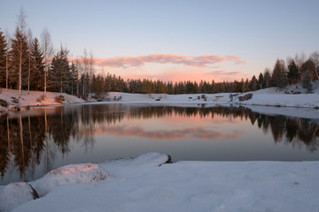 Fototapeta na wymiar Winter landscape. The forest lake is partially covered with ice, surrounded by rocks, shrubs and spruces against the evening sky painted by the rays of the setting sun.
