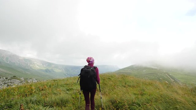 Tourist woman Nordic walking with sticks and a large backpack among the grass in the Caucasus on a high mountain in windy weather in the fall, Adygea, Russia. Sporty woman hiking in autumn, back view.