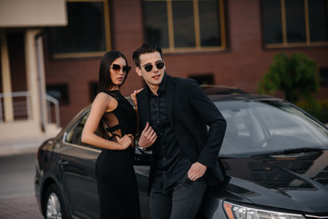 Fototapeta na wymiar A young stylish couple in black stands near the car at sunset. Fashion and style