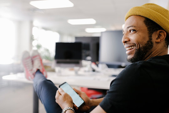 Happy Businessman Using Smart Phone With Feet Up In Office