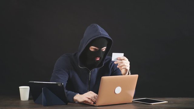 Computer connoisseur man hacking laptop system, want to hack bank card, sit in hood of pullover and black balaclava mask, inspires fear sitting in the dark room, black background