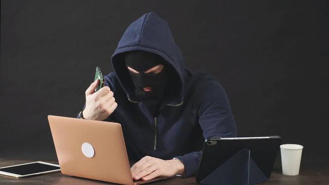 Male hacker in black mask and hood uses laptop to hack a hard drive while in dark office room.