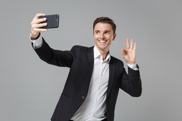 Smiling young business man in classic black suit shirt isolated on grey background. Achievement career wealth business concept. Mock up copy space Doing selfie shot on mobile phone showing OK gesture.