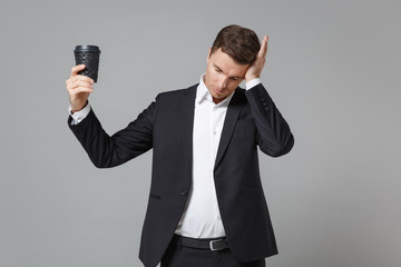 Naklejka premium Tired young business man in classic black suit shirt posing isolated on grey background. Achievement career wealth business concept. Mock up copy space. Hold cup of coffee or tea, put hand on head.