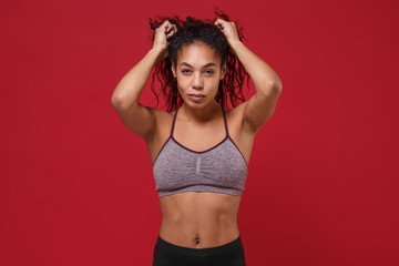 Obraz premium Beautiful young african american sports fitness woman in sportswear posing working out isolated on red wall background studio portrait. Sport exercises healthy lifestyle concept. Put hands on head.