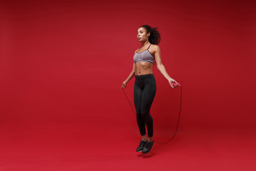 Strong young african american sports fitness woman in sportswear posing working out isolated on red background studio portrait. Sport exercises healthy lifestyle concept. Jumping on skipping rope.