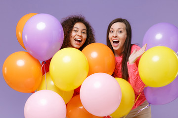 Fototapeta na wymiar Excited two european african american women friends in knitted sweaters isolated on violet purple background. Birthday holiday party, people emotions concept. Celebrating hold colorful air balloons.