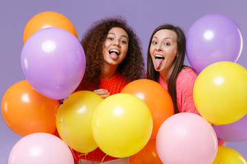 Fototapeta na wymiar Funny two european african american women friends in knitted sweaters isolated on violet purple wall background. Birthday holiday party concept. Celebrating hold colorful air balloons showing tongue.