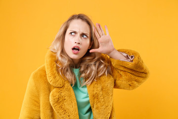 Shocked young blonde woman girl in yellow fur coat posing isolated on orange wall background studio portrait. People emotions lifestyle concept. Mock up copy space. Try to hear you with hand near ear.