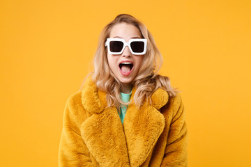 Surprised young blonde woman girl in yellow fur coat, dark sunglasses posing isolated on orange...