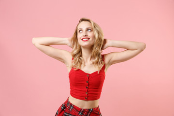 Pensive smiling young blonde woman girl in red sexy clothes posing isolated on pastel pink background in studio. People lifestyle concept. Mock up copy space. Holding hands behind head, looking up.