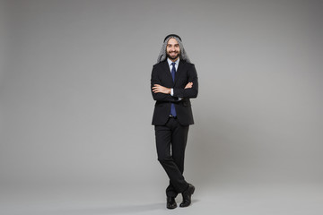 Smiling bearded young arabian muslim businessman in keffiyeh kafiya ring igal agal black suit shirt tie isolated on gray background. Achievement career wealth business concept. Holding hands crossed.