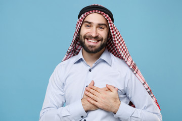 Smiling young bearded arabian muslim man in keffiyeh kafiya ring igal agal casual clothes isolated on pastel blue background. People religious lifestyle concept. Holding hands folded on heart, chest.